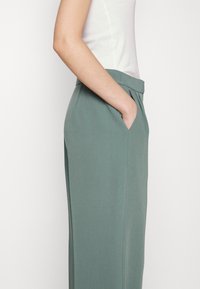 Pantalons vert clair avec une coupe ajustée, comprenant des poches latérales, un devant plat et une texture lisse. Conçus pour une silhouette élancée.