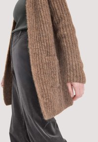 Cardigan en tricot marron avec un motif texturé, doté de manches longues et d'une poche frontale, associé à un pantalon en velours côtelé noir.