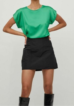 Blouse courte en satin vert à manches courtes avec une coupe décontractée, associée à une jupe mini noire moulante. Le mannequin se tient avec les mains sur les hanches, portant des bottes mi-hautes.