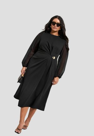 Frau trägt ein schwarzes Midikleid mit transparenten langen Ärmeln, goldfarbenem Gürtel, schwarzen Riemchenpumps, Sonnenbrille und hält eine kleine schwarze Handtasche.