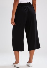 Svarta culotter i lätt tyg, med elastisk midja. Nederkanterna når mitt på vaden, matchas med vita slip-on skor.