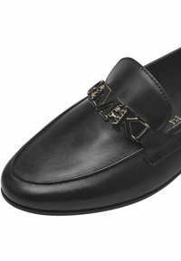 Zwarte leren loafers met een platte zool en een opvallend metallic logo accent aan de voorkant. Glad textuur met gestikte details.
