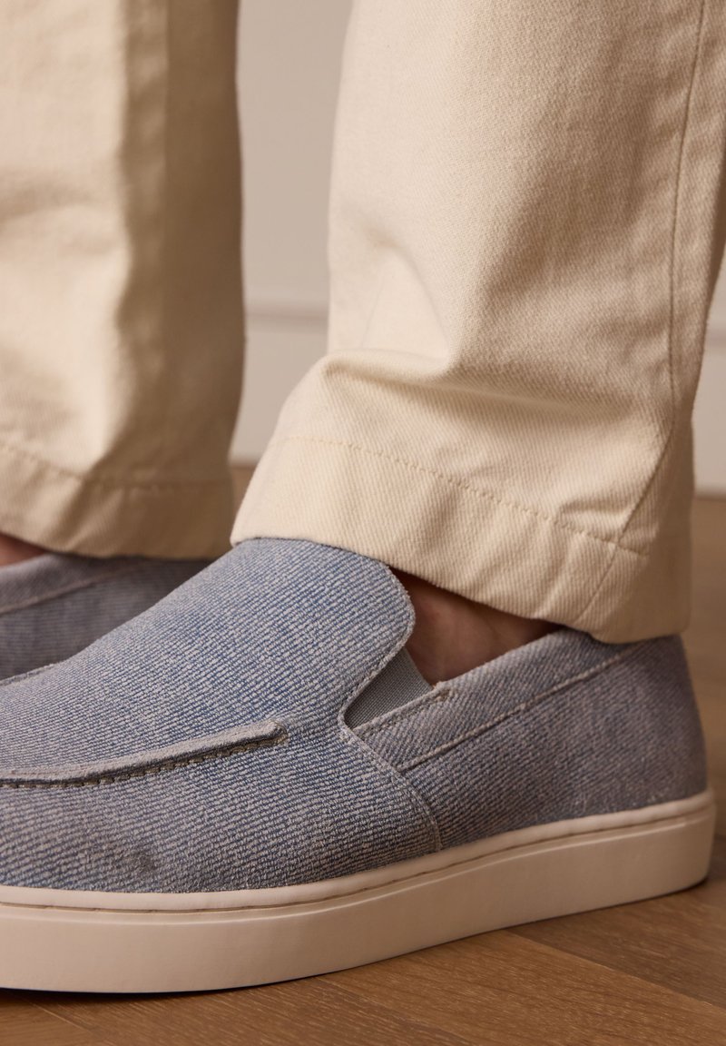 Blauwe denim slip-on schoenen met een gestructureerde oppervlakte en elastische details, gecombineerd met lichtbeige broek, tegen een houten vloer.