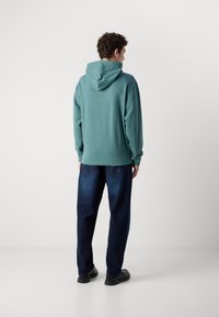 Felpa con cappuccio color turchese, dal taglio rilassato, con polsini e orlo a coste, abbinata a jeans blu scuro a vestibilità ampia. Design minimalista, superficie liscia.