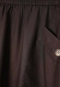 Gros plan sur une jupe en tissu marron foncé avec une ceinture élastique et une poche à rabat sécurisée par un bouton rond à deux trous.