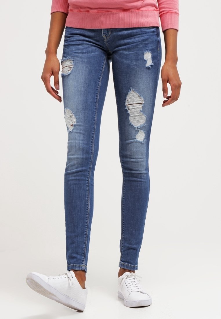 Jeans ajustados en denim azul medio, con parches desgastados en ambas piernas. Combinados con zapatillas de lona blancas para un look casual.