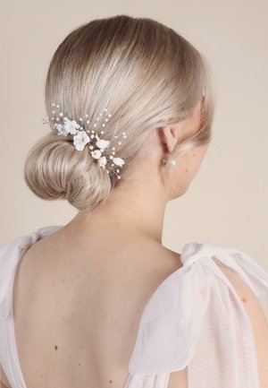 Fibbia floreale bianca con accentuazioni di perle, sistemata in un basso chignon. Presenta fiori e rami delicati, in contrasto con capelli chiari e ondulati.