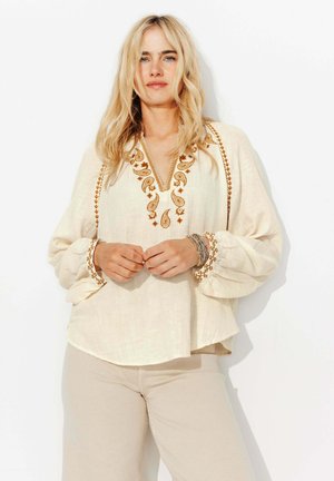 Femme blonde portant un chemisier crème avec des broderies marron au niveau du col et des poignets, un pantalon beige et des bracelets empilés, se tenant devant un fond blanc.