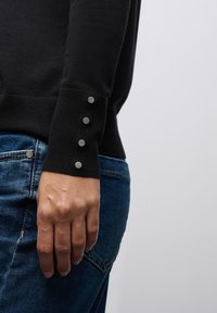 Maglione nero con polsini a coste e quattro bottoni a pressione in metallo, abbinato a jeans in denim blu. Design semplice e forma aderente.