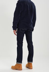 Granatowa polarowa kurtka z teksturowanym wykończeniem, w zestawieniu z ciemnoniebieskimi dżinsami i tanowymi butami roboczymi. Ubranie ma luźny krój i funkcjonalny design.