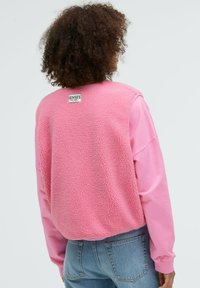 Rosa Fleece-Sweatshirt mit einer weichen Textur, mit einem Etikett auf der Rückseite. Lange Ärmel und ein lockerer Schnitt. Kombiniert mit blauen Denim-Jeans.