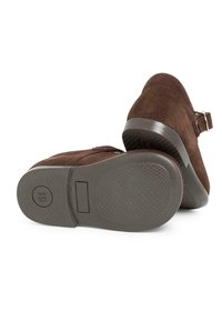 Braune Wildlederschuhe mit Riemen und Schnalle, ausgestattet mit einer strukturierten Gummisohle und einer abgerundeten Spitze. Die Rückansicht zeigt eine glatte Ferse mit Akzent.