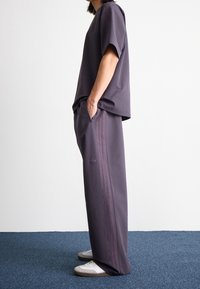 Fialový sportovní set se skládá z měkkého, lehkého trika s krátkým rukávem a odpovídajících kalhot s bočními pruhy a logovými akcenty, doplněný o bílé tenisky.