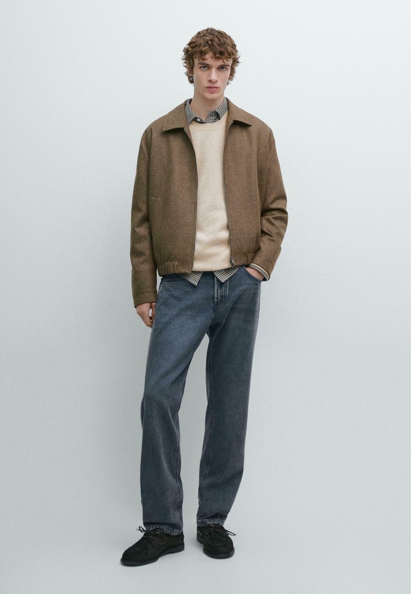 Bruine jas van wolmix over een beige trui, gecombineerd met donkere spijkerbroeken. Voorzien van een geruit overhemd en zwarte schoenen.