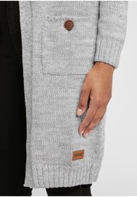 Gray knitted cardigan made of soft material, featuring a front pocket, a brown button closure, and a small brand tag at the hem.