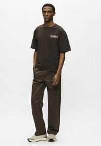 Joven de pie vistiendo una camiseta oversized de color marrón oscuro con el logo "Double", pantalones marrones a juego y zapatillas deportivas blancas sobre un fondo blanco.