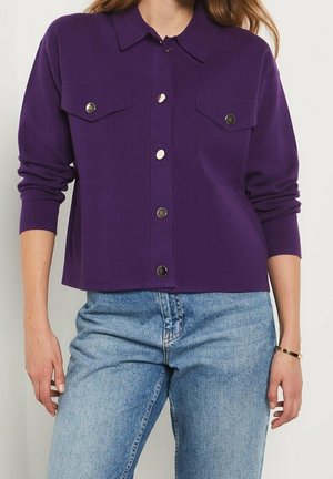 Femme portant une chemise violette à manches longues avec des poches poitrine et un jean bleu clair, debout les bras détendus le long du corps.