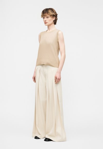 Γυναίκα με αμάνικη μπεζ μπλούζα και ευρύχωρο, εκρού παντελόνι ψηλόμεσο, στέκεται με ουδέτερη έκφραση σε απλό λευκό φόντο.