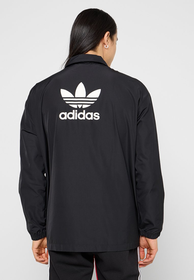 Schwarze Windbreaker-Jacke aus glattem Material mit einem weißen Adidas-Logo und drei Streifen auf dem Rücken. Lange Ärmel mit elastischen Bündchen.