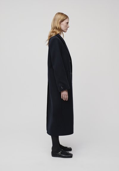 Long navy coat made of thick fabric, featuring a notched collar, two front pockets, and button closures. Worn with black shoes and tights.