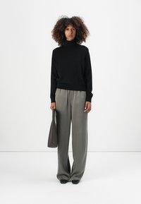 Pull à col roulé noir, pantalon gris ample, tenant un sac à main gris, sur fond blanc. Le pull a des manches longues ; le pantalon est à jambe large.