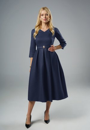 Blondynka w granatowej, midi sukience z paskiem, z rękawami do łokci, w czarnych szpilkach, stojąca na szarym tle.