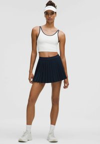 Bijeli crop top s tamnoplavim detaljima, uparen s tamnoplavom plisiranom suknjom. Model nosi bijele tenisice i vizir. Glatka tkanina s prilagođenim krojem.