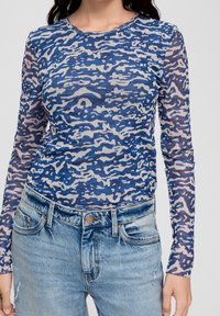 Kvinna iklädd en långärmad topp med abstrakt mönster i blått och beige, tillsammans med ljusblå högmidjade jeans.