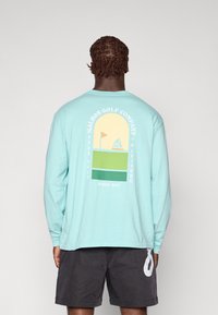 Lys blå langermet skjorte med grafisk design på ryggen som har en seilbåt, flagg og fargede blokker i gult og grønt.