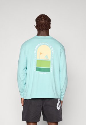 Lys blå langermet skjorte med grafisk design på ryggen som har en seilbåt, flagg og fargede blokker i gult og grønt.