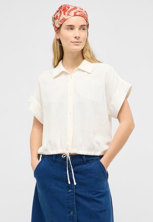 Kvinde iført en hvid kortærmet skjorte med knapper og snøre i taljen, blå denimnederdel og rødt mønstret tørklæde, hænderne i lommerne.