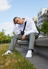 Hvid hættebluse med sort logo, grøn cargo-bukser og hvide sneakers med sorte accenter. Sidder på en grå stenskrænt i et græsareal.