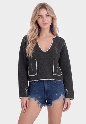 Blond kvinde iført en mørkegrå V-hals sweater med hvid kant og blomsterbroderi, kombineret med flossede blå denimshorts og blå kvastøreringe.