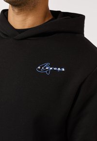 Mann trägt einen schwarzen Hoodie mit einem blauen, kursiv gedruckten "Closure"-Logo auf der linken Brusttasche.