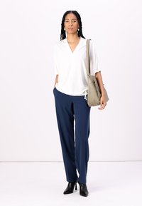 Witte blouse met korte mouwen en V-hals, gecombineerd met marineblauwe broek. Het model draagt een lichtgrijze schoudertas en heeft zwarte haklaarzen aan.