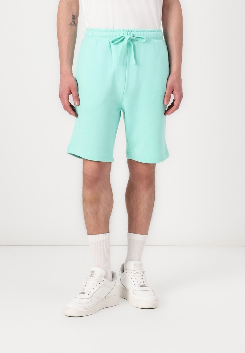 Homme portant un short à cordon de serrage vert menthe, des chaussettes blanches montantes et des baskets blanches, debout devant un fond blanc uni.