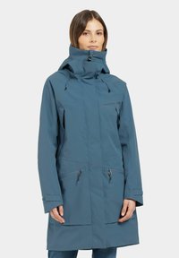 Veste imperméable bleue avec col montant, capuche ajustable, poches zippées sur la poitrine et sur les côtés, et une coupe légèrement décontractée. Texture lisse.