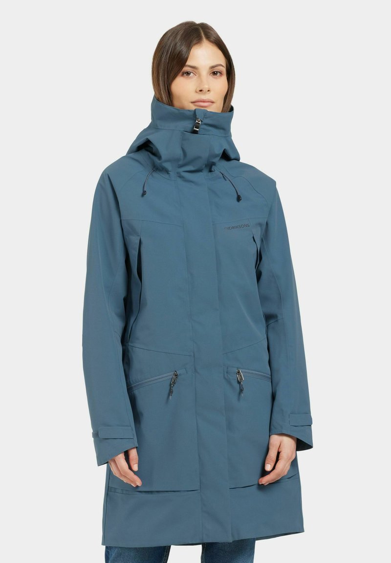 Veste imperméable bleue avec col montant, capuche ajustable, poches zippées sur la poitrine et sur les côtés, et une coupe légèrement décontractée. Texture lisse.