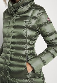 Padded green coat with high collar, featuring quilted texture, side pockets, and logo on the sleeve. Made from glossy, lightweight material.