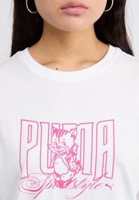 Bílá bavlněná tričko s velkým růžovým grafikou kreslené kočky. Design obsahuje tučné texty "PUMA" a hravé písmo.