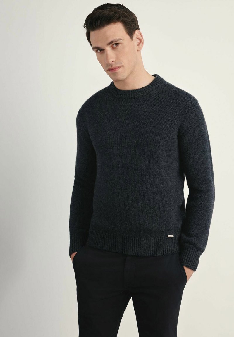 Granatowy sweter z dzianiny z prążkowanym okrągłym dekoltem, teksturowanym materiałem i małym metalowym logo na dolnym brzegu. Nosi się go z ciemnymi spodniami.