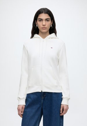 Γυναίκα με μακριά σκούρα μαλλιά φοράει λευκό φούτερ με φερμουάρ και μικρό λογότυπο καθώς και μπλε τζιν, στέκεται μπροστά σε απλό φωτεινό φόντο.