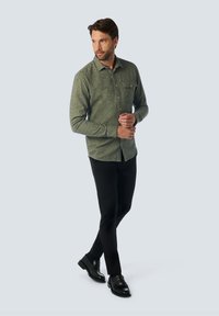 Groen gestippeld overhemd met een borstzak, gecombineerd met zwarte slim-fit broek en zwarte schoenen, staand tegen een lichte achtergrond.