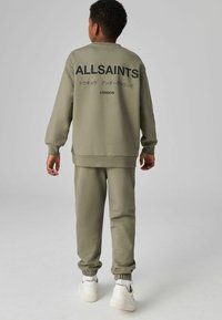 Oliņu zaļa sporta krekla un bikses komplekts ar ribu aprocēm, aizmugurē ar "ALLSAINTS" logo. Modelis valkā baltas kedas.