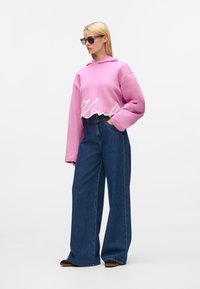 Rosa, taillierte Kapuzenjacke mit "Karl" in weißer, geschwungener Schrift am Saum. Kombiniert mit hochgeschnittenen, weiten blauen Jeans und schwarzen Slippern.