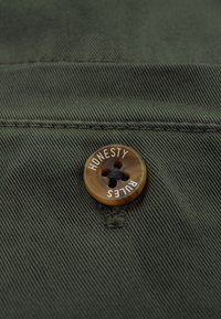 Olivengrüner Stoff mit einem braunen Knopf, der mit dem eingeprägten Text "EHRLICHKEIT REGIERT" und einem Vierlochdesign versehen ist, wodurch die Textur des Knopfes hervorgehoben wird.