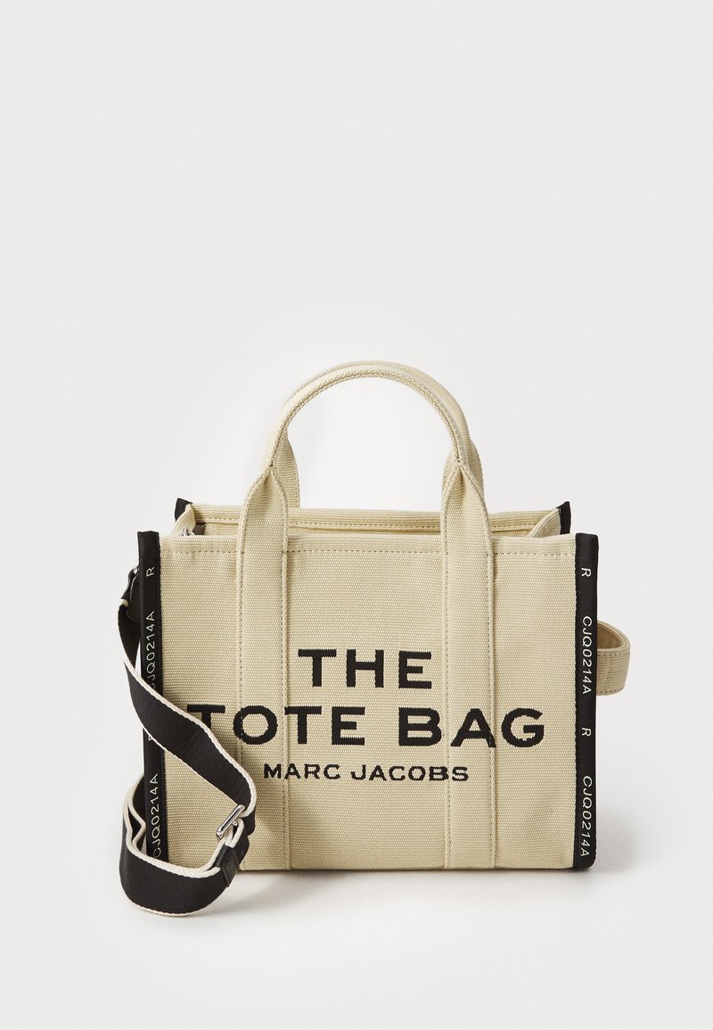 Beige canvas tote-väska med svart text och accenter, dubbla topphandtag och en justerbar rem. Har en rektangulär form och minimalistisk design.