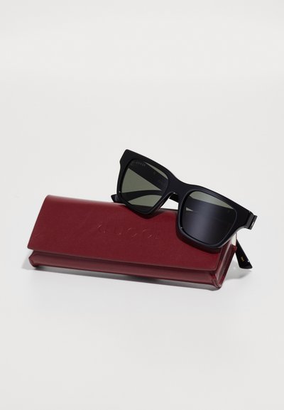 Ochelari de soare rectangulari negri, cu lentile întunecate, așezați pe un toc din piele roșie, împodobit cu "GUCCI." Textură netedă și design modern.