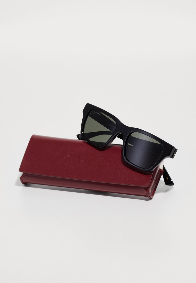 Black rectangular sunglasses with dark lenses, resting on a red leather case embossed with "GUCCI." Smooth texture and modern design.