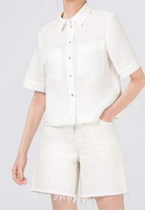 Femme portant une chemise blanche à manches courtes avec deux poches poitrine et un short en denim blanc à ourlet effiloché.